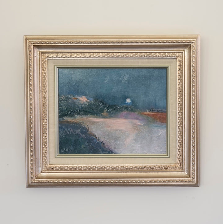 A framed landscape painting featuring a dark blue night sky, distant mountains, and a pale glowing moon over a soft-toned foreground, housed in an ornate gold-toned frame mounted on a light beige wall.
